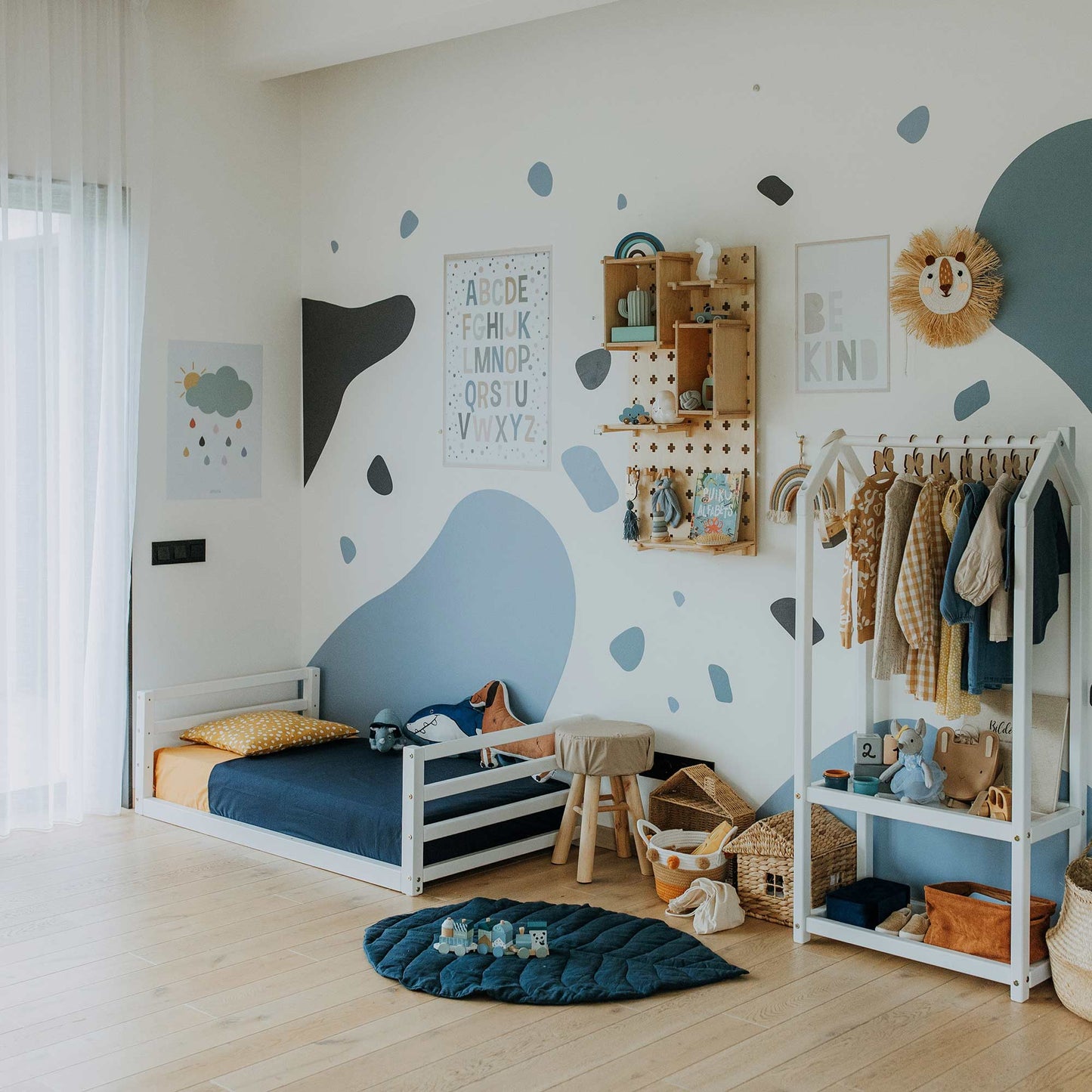 A children's bedroom with a blue-themed decor, featuring a toddler floor bed with a horizontal rail headboard and footboard, wall shelves, a small wardrobe filled with clothes, and various toys. The walls boast playful blue and white patterns along with educational posters. This room is the perfect transitional space from toddler to big kid.