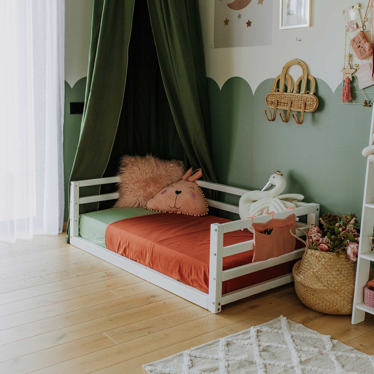 A children's room with a white floor-level bed equipped with a 3-sided safety rail, crafted from solid pine. The bed features vibrant orange bedding and is surrounded by stuffed animals. Green curtains frame the window, while playful wall decor adds charm to the space. A wicker basket filled with flowers rests on the floor beside the bed, ensuring a cozy and delightful atmosphere for little ones.