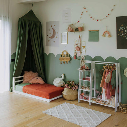 A cozy child's bedroom features a green canopy toddler floor bed with a horizontal rail headboard and a solid wood frame, playful wall decor, and a clothing rack with hanging garments. The room has light wooden flooring, a patterned rug, and various soft toys and decorations to encourage independent sleeping.