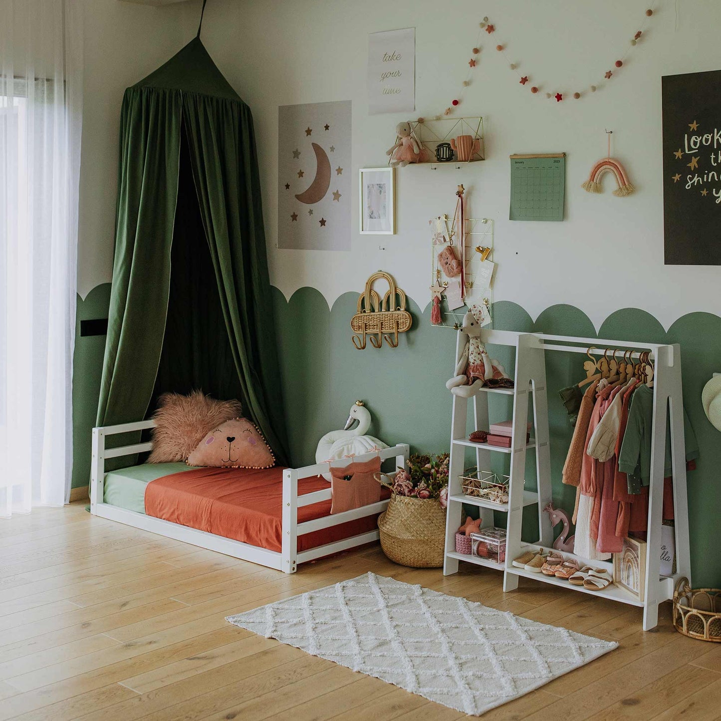 A stylish children's room featuring a Toddler floor bed with a horizontal rail headboard and footboard under a canopy, complete with wall decorations, a small clothes rack with hanging garments, and a white rug on a solid wood floor. This transitional piece combines elegance and practicality for your little one's space.