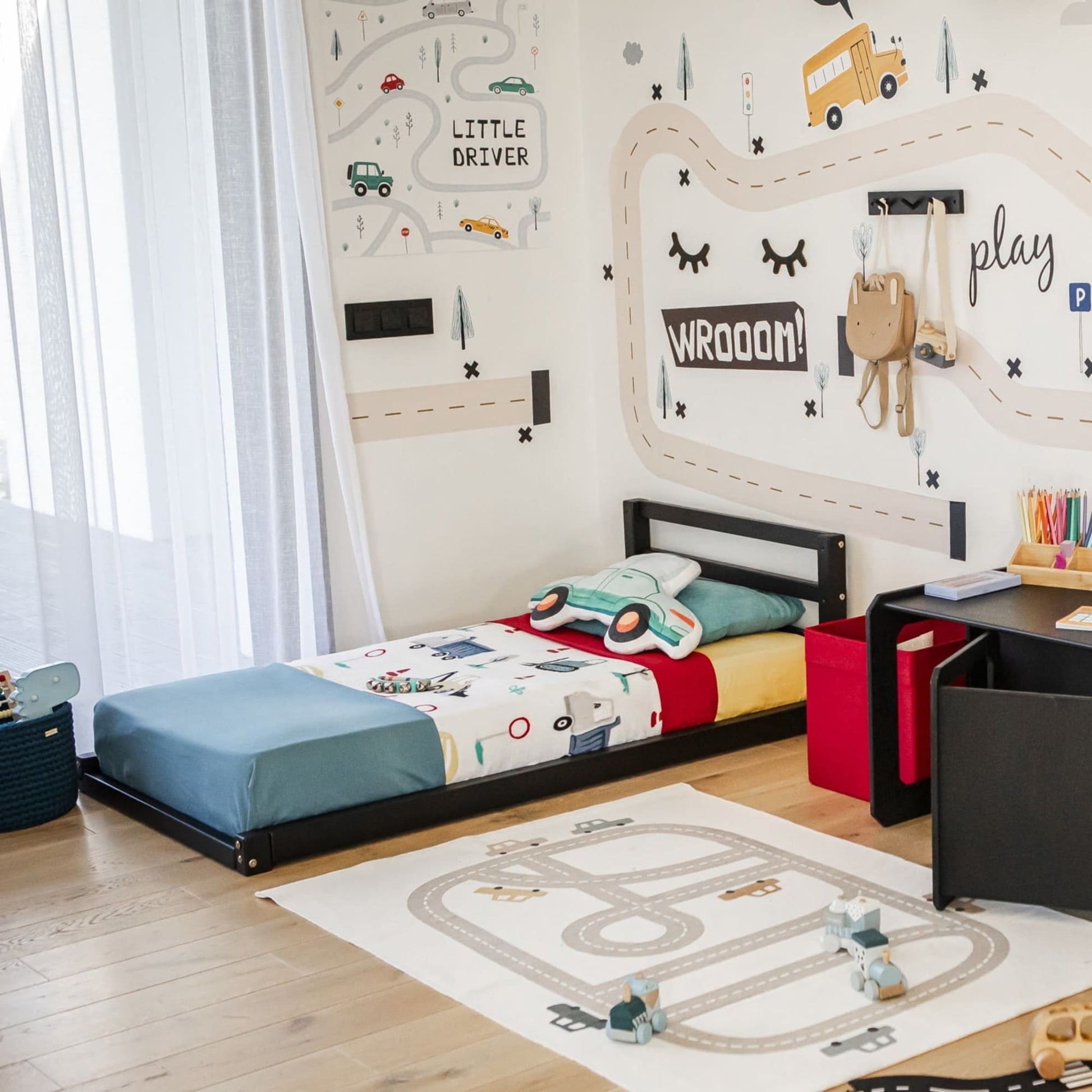 A child's bedroom featuring a car-themed wall decoration, a low bed with colorful bedding on a Toddler floor bed with a horizontal rail headboard, various toys, and a desk with craft supplies. There is a road-patterned rug on the wooden floor.