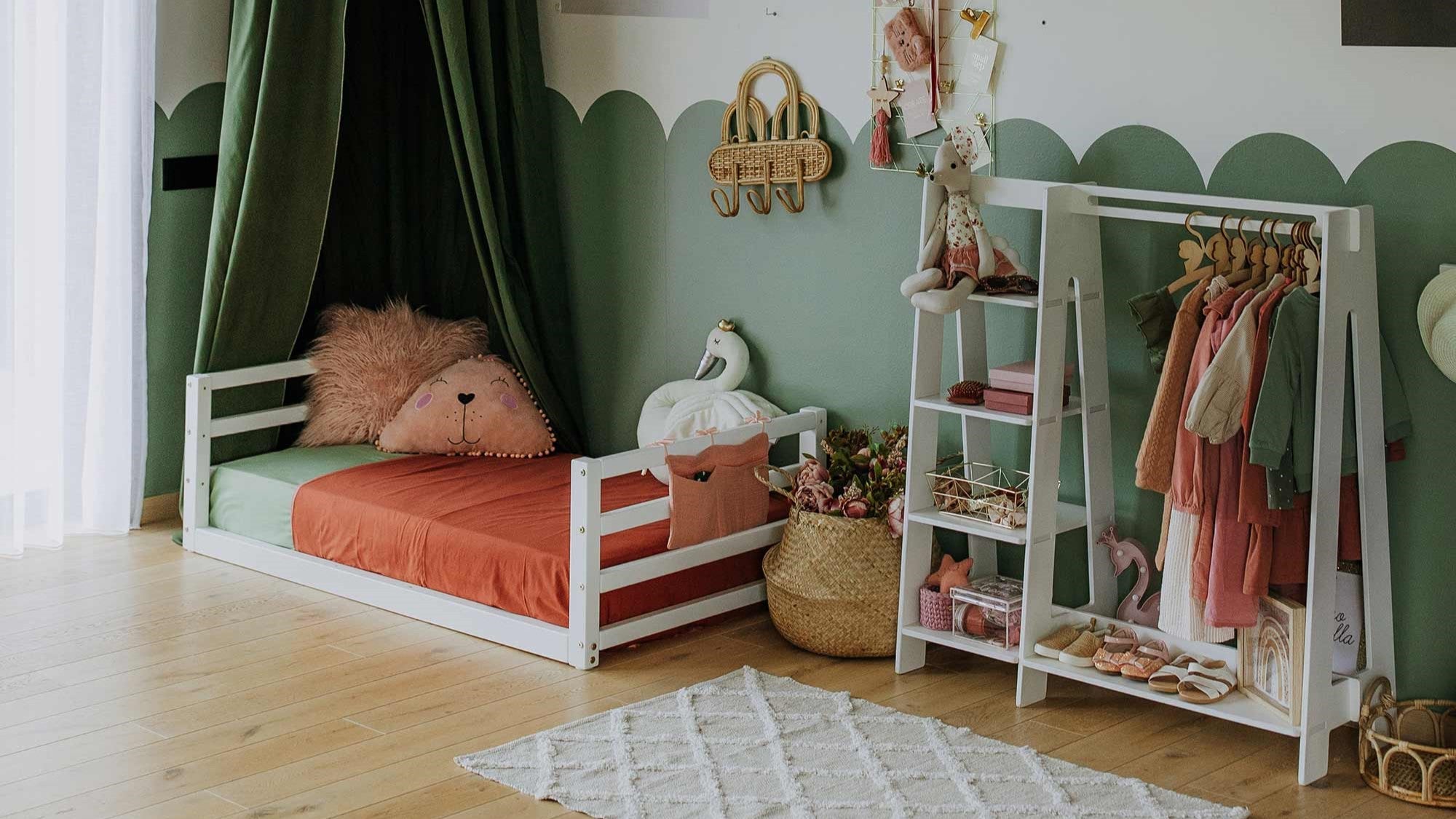 A children's bedroom with a toddler floor bed featuring a horizontal rail headboard and footboard, plush toys, a clothing rack, and decorative items. The room showcases green and white walls, wooden flooring, and a white patterned rug—perfect for encouraging independent sleeping.