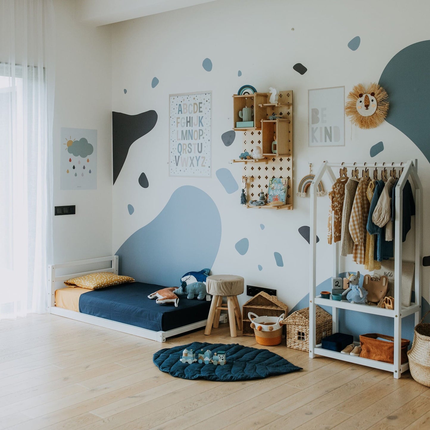 A children's bedroom featuring a solid wood frame bed, specifically the Toddler Floor Bed with a Horizontal Rail Headboard, along with a bookshelf, clothing rack, toys, and wall decor in blue and white. There's a ladder-style shelf on the right and various storage baskets on the wooden floor, creating an inviting space for independent sleeping.