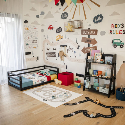 A children's bedroom with a car-themed decor, featuring a Children's Floor Level Bed with a 3-Sided Safety Rail, shelves full of toys, and wall stickers of vehicles and road signs. The room boasts hardwood flooring and is illuminated by natural light from a window adorned with white curtains.