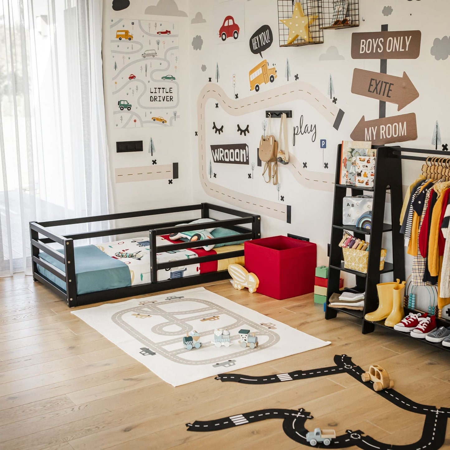 A children's room with a car-themed decor, featuring a floor-level kids' bed with a horizontal rail fence, wall decals, toy storage, clothes rack, and a play mat with toy cars on a wooden floor. This setup encourages independent sleeping in style.