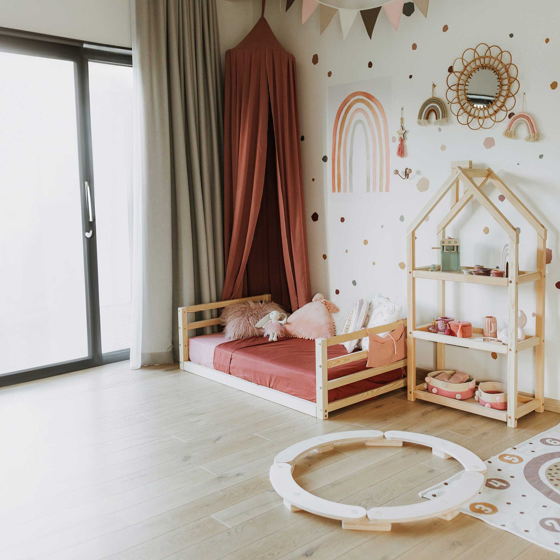 A children's room featuring a toddler floor bed with a horizontal rail headboard and footboard, complete with a solid wood frame, canopy, and wood shelf. Decor includes a rainbow wall hanging, polka dot wallpaper, and a play area with a circular wooden track encouraging independent sleeping.