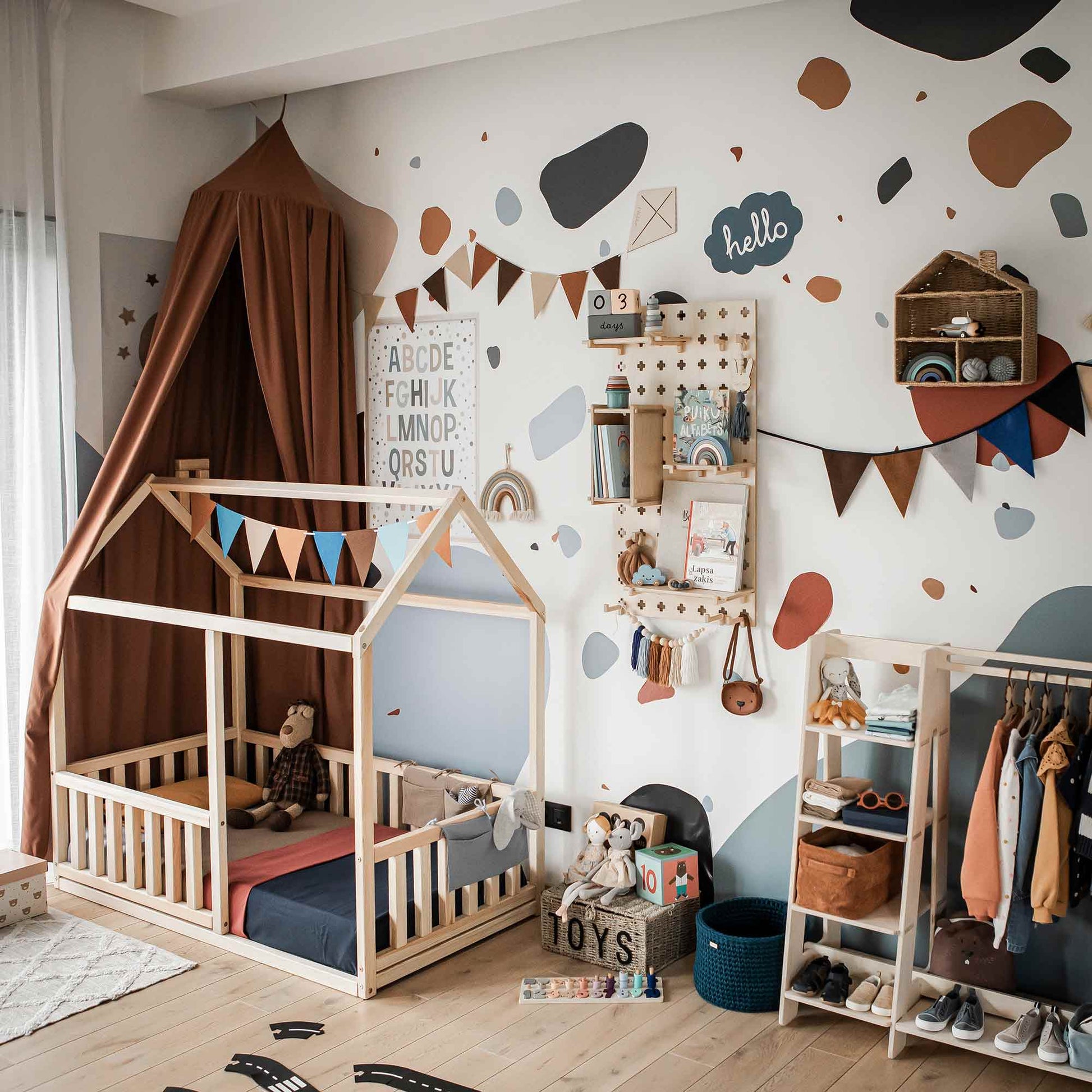 A well-organized children's bedroom features a Montessori floor house bed with rails, a wall-mounted alphabet chart, shelves filled with toys and decor, and neatly hung clothes. The walls are adorned with abstract patterns and colorful banners.