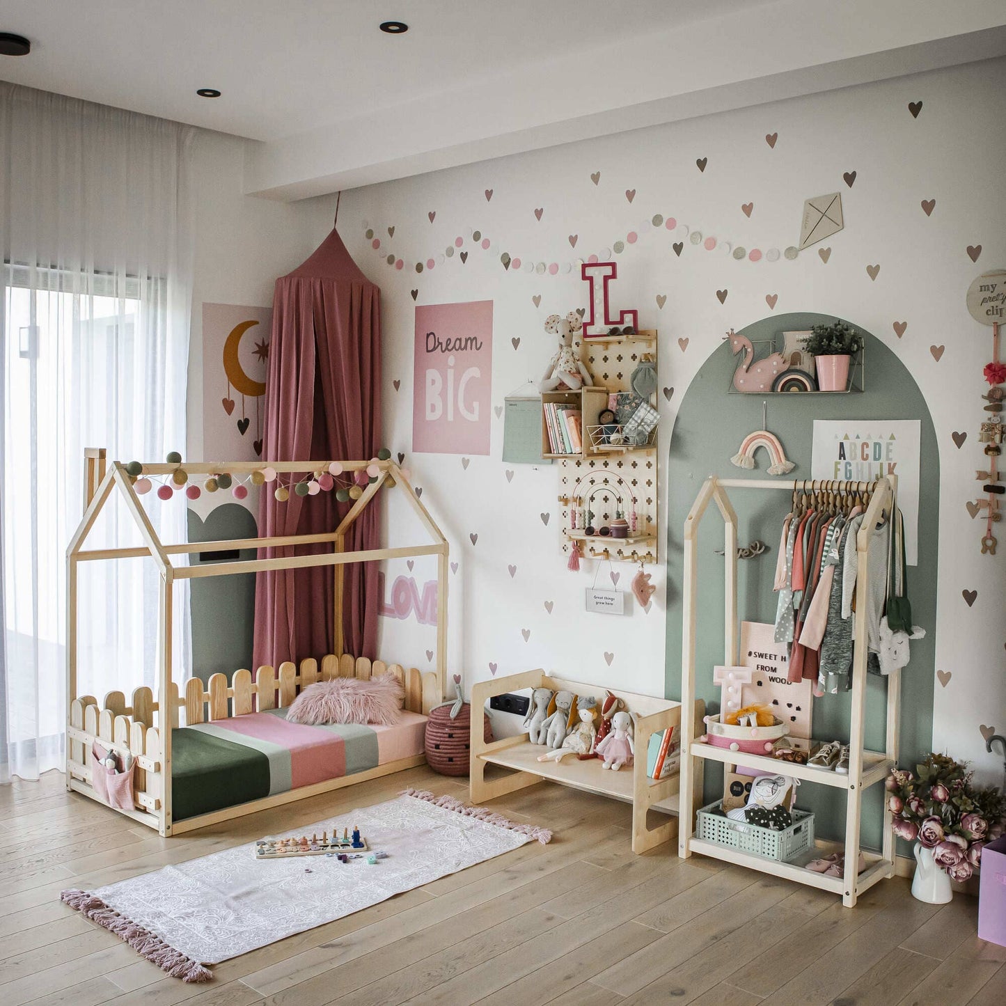 A child's room featuring a playhouse bed, a reading bench, a Montessori wardrobe, and shelves holding toys and decor. The walls are adorned with a "Dream Big" sign, hearts, and various pastel murals, all reflecting Montessori principles to inspire independence and creativity.