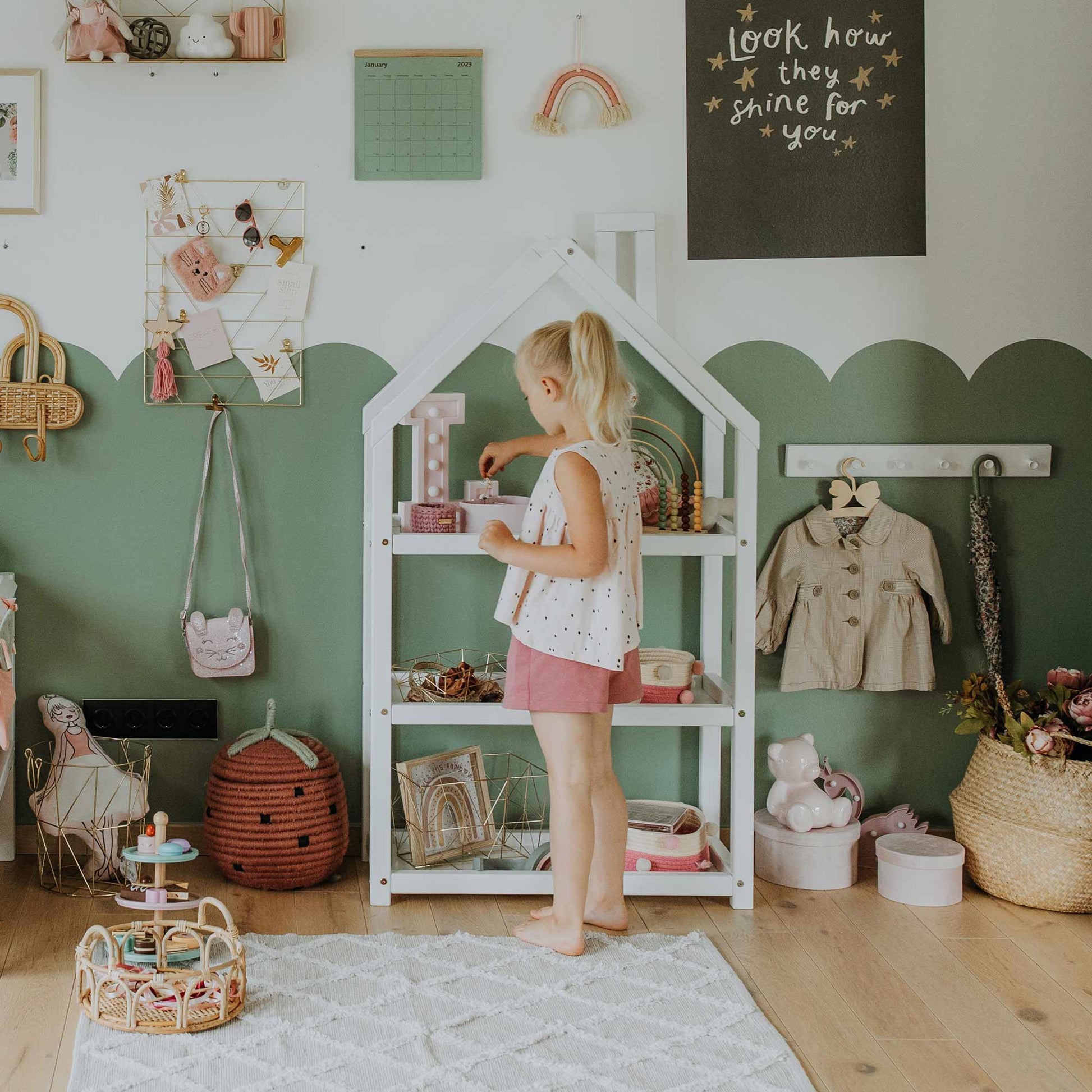 A child in a decorated room stands beside a House-shaped Montessori shelf. The room features green and white walls, a variety of toys, and a "Look how they shine for you" sign.