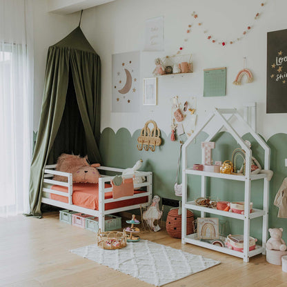 A children's room with a small bed draped in a canopy, featuring a house-shaped Montessori shelf filled with toys and books. The walls are adorned with pictures and garlands, set against a light wooden floor, and a large window with sheer curtains completes the cozy space.