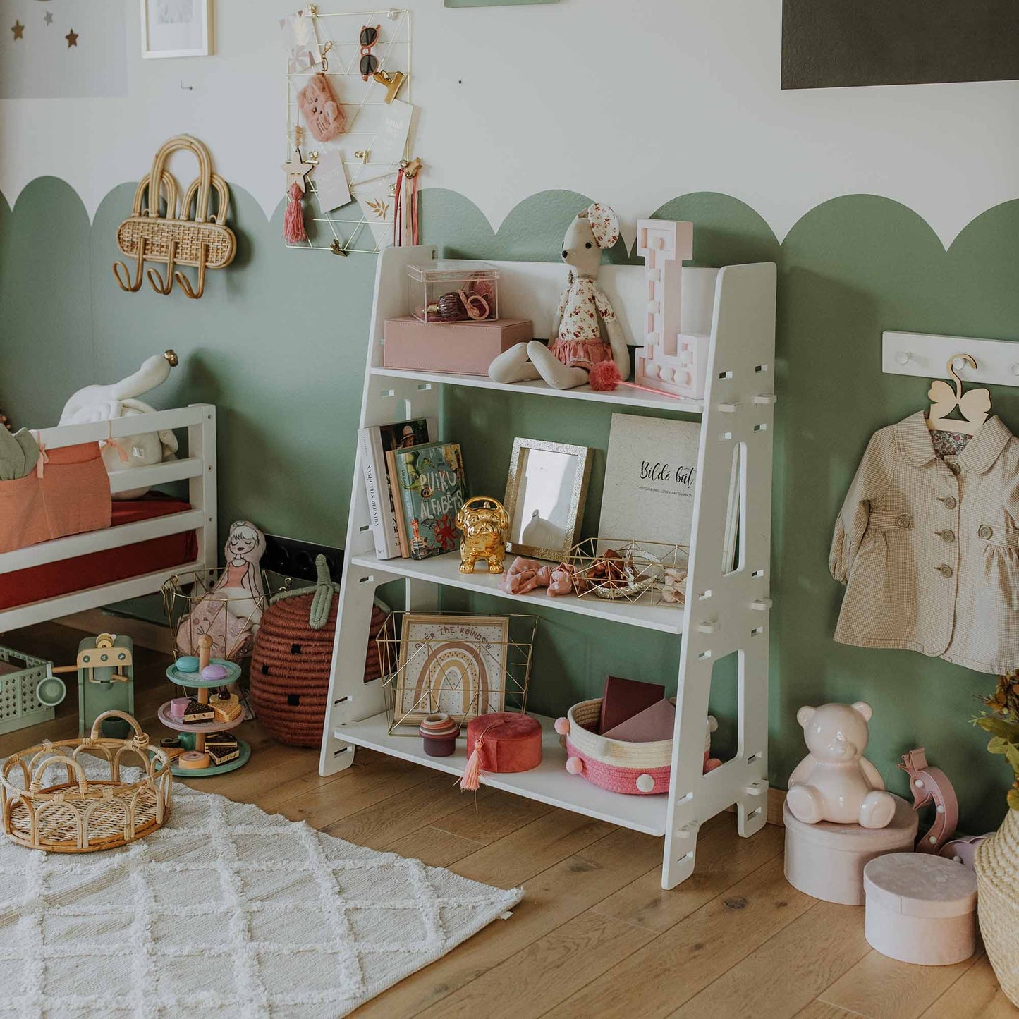 A nursery with green and white walls features a Montessori toy shelf holding toys and knick-knacks. A small bed is visible on the left, while accessories and clothing hang neatly on the wall. A cozy rug lies on the wooden floor.