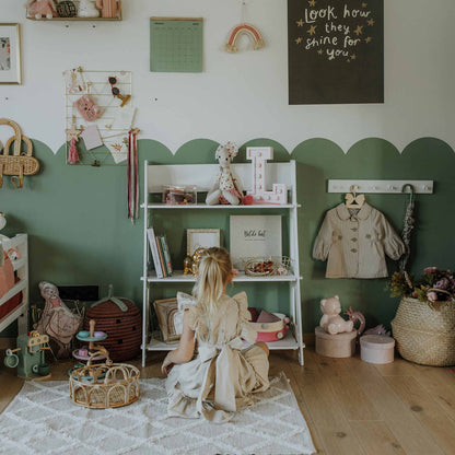 A young child sits on a rug in a neatly organized nursery, surrounded by toys, books, and decorative items neatly arranged on a Montessori toy shelf against walls painted green and white.