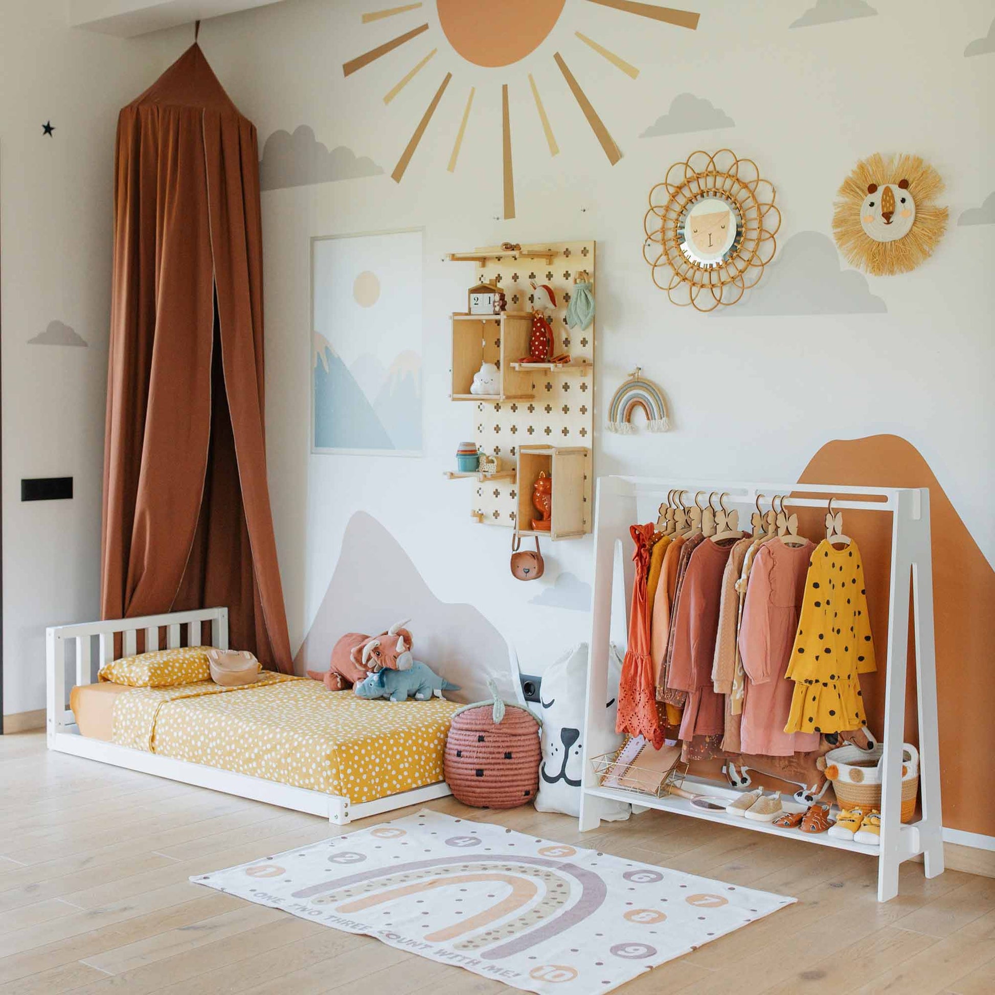 A children's room showcasing a low bed, a play rug, the Kids' clothing rack brimming with colorful outfits, a mural on the wall, and various decorations. The overall theme features warm tones and nature-inspired designs.