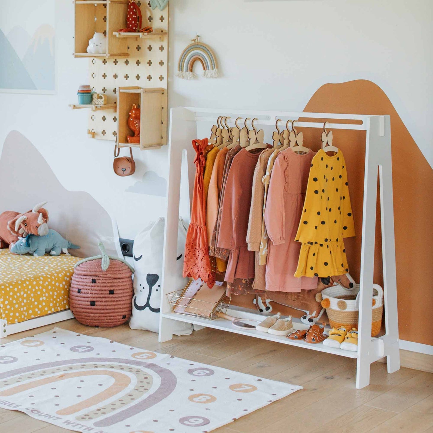 A neatly organized children's room features a Kids' clothing rack displaying colorful garments, a toy storage basket, a small bookshelf, and a decorative rainbow rug on the floor.