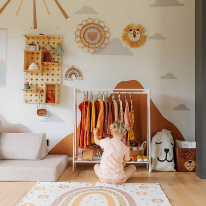 A child sits on the floor in a pastel-themed room, surrounded by a Kids' clothing rack, whimsical wall decorations, and a plush lion bag.