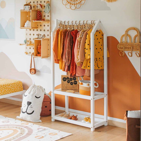 A children's room featuring a Montessori wardrobe displaying colorful outfits. A plush animal-shaped pillow, hanging shelves adorned with decorations, and pastel-themed wall decor are also visible.