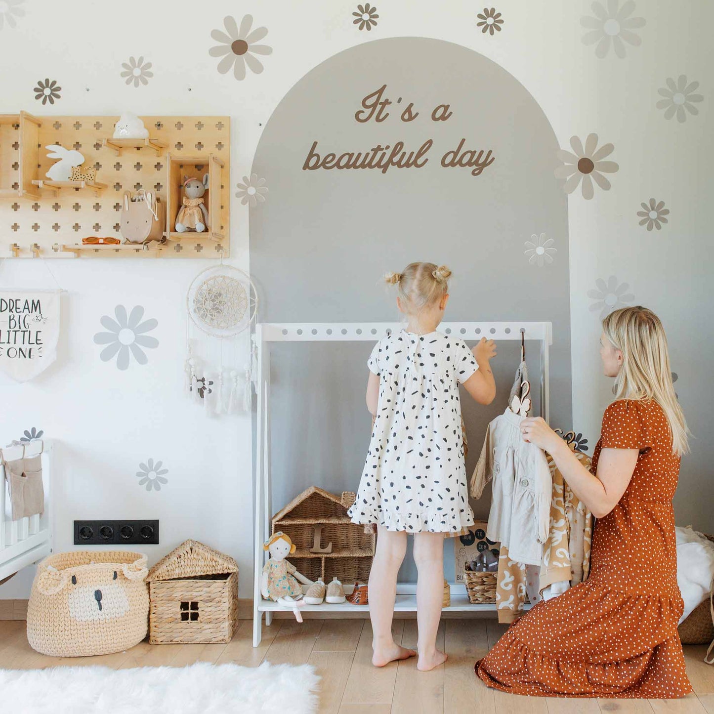 A woman and a child organize a dollhouse and toys in a room decorated with floral wall decals and text saying "It's a beautiful day." The woman is wearing a rust-colored dress, and the child is dressed in polka dots. In the corner, an open A-frame kids' clothing rack showcases their neatly arranged outfits.