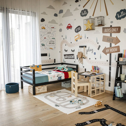 A child's bedroom with a car theme, featuring a bed, a 2-in-1 table and chair set or activity cube, various toys including a toddler toy set, and educational books. The walls are decorated with car-themed murals and motivational signs.