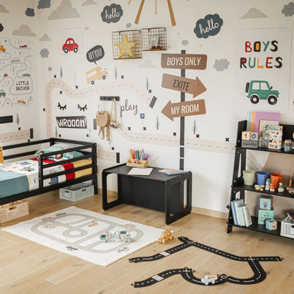 A children's room with a road-themed décor, featuring wall stickers of roads and traffic signs, a bed, a Montessori weaning table for self-feeding or playtime, a desk with supplies, a play mat with a road print, and shelves filled with books and toys.