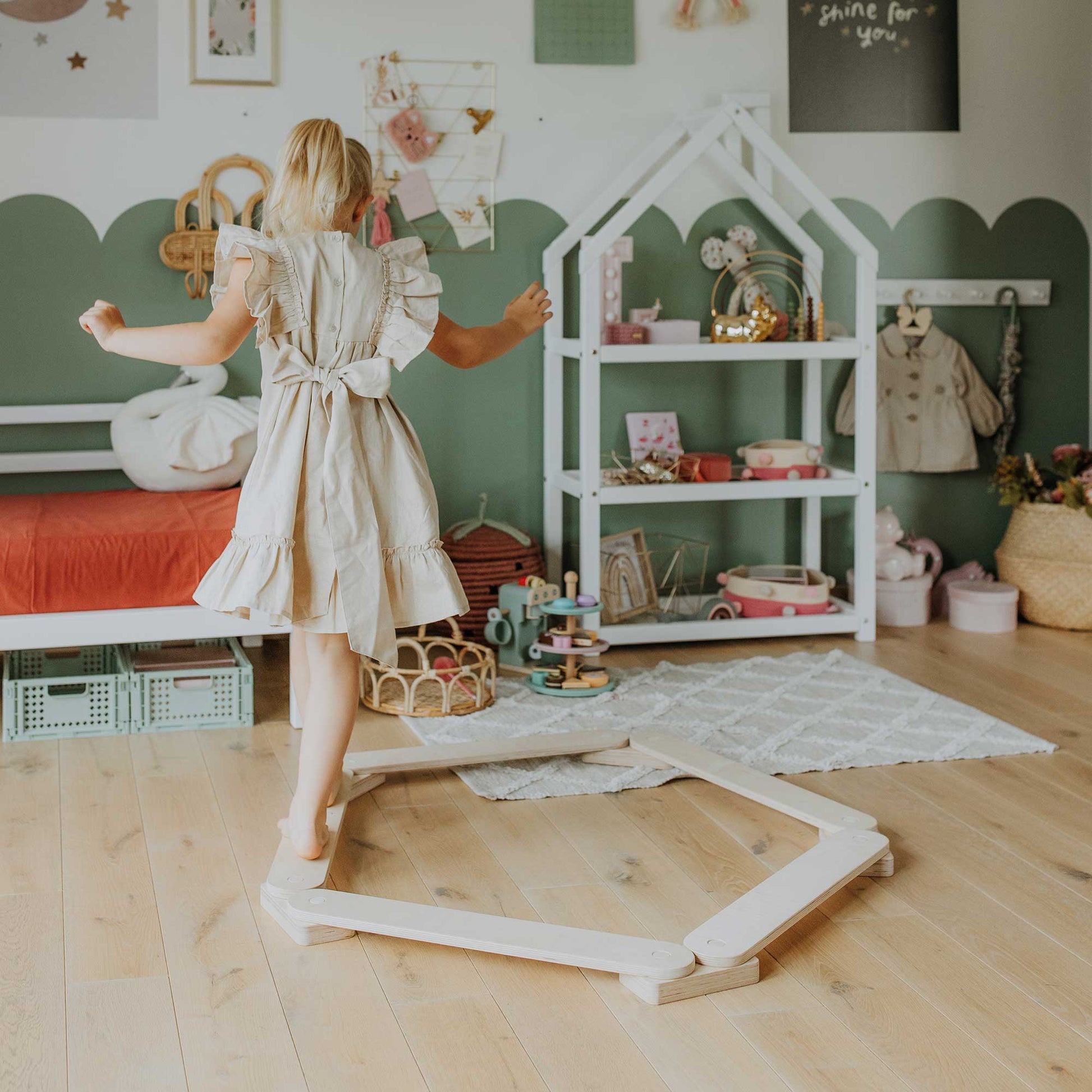 A young girl in a beige dress confidently stands on a Balance Beam Set in a beautifully decorated playroom filled with Montessori toys, enhancing her motor skills amidst the various playthings, a small bed, and charming wall decor.