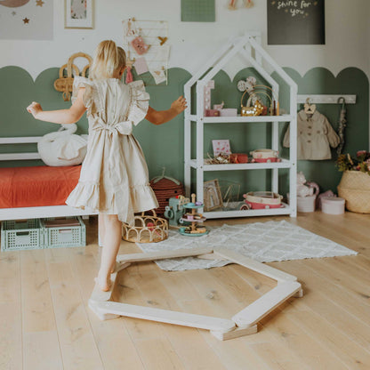 A young girl in a beige dress confidently stands on a Balance Beam Set in a beautifully decorated playroom filled with Montessori toys, enhancing her motor skills amidst the various playthings, a small bed, and charming wall decor.