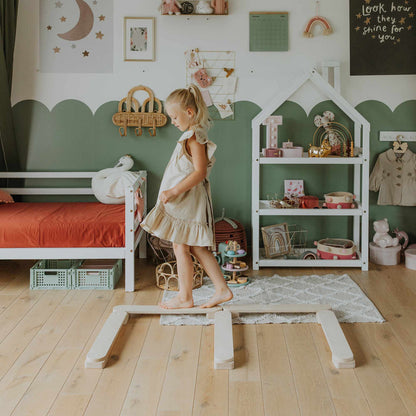 A young girl in a dress balances on a wooden Balance Beam Set in a child's bedroom with green and white walls, shelves, and toys, enhancing her motor skills like a Montessori toy.