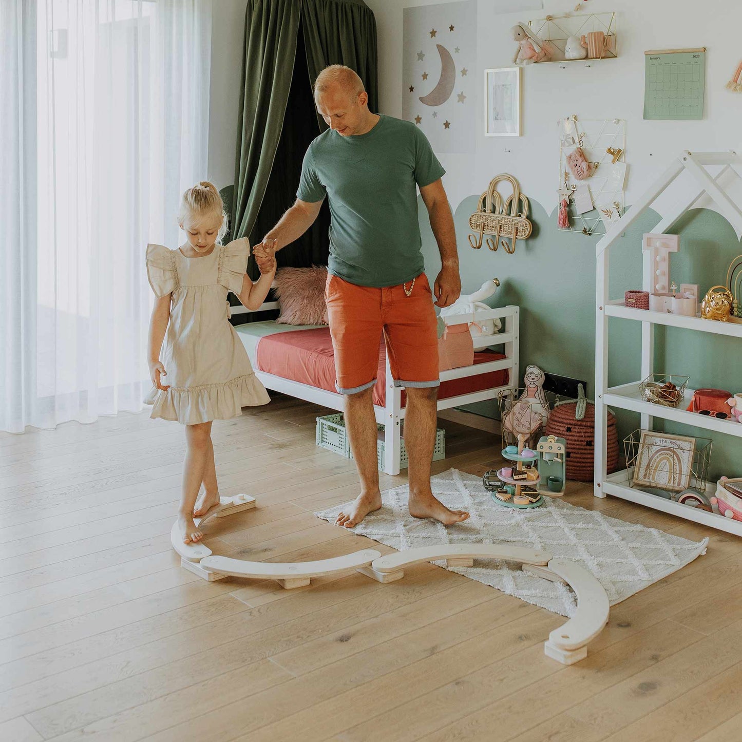 A man with a young girl, holding her hand, is walking barefoot on a curved round balance beam in a playful, well-decorated bedroom. This Montessori indoor activity toy enhances their motor skills while creating joyful moments together.