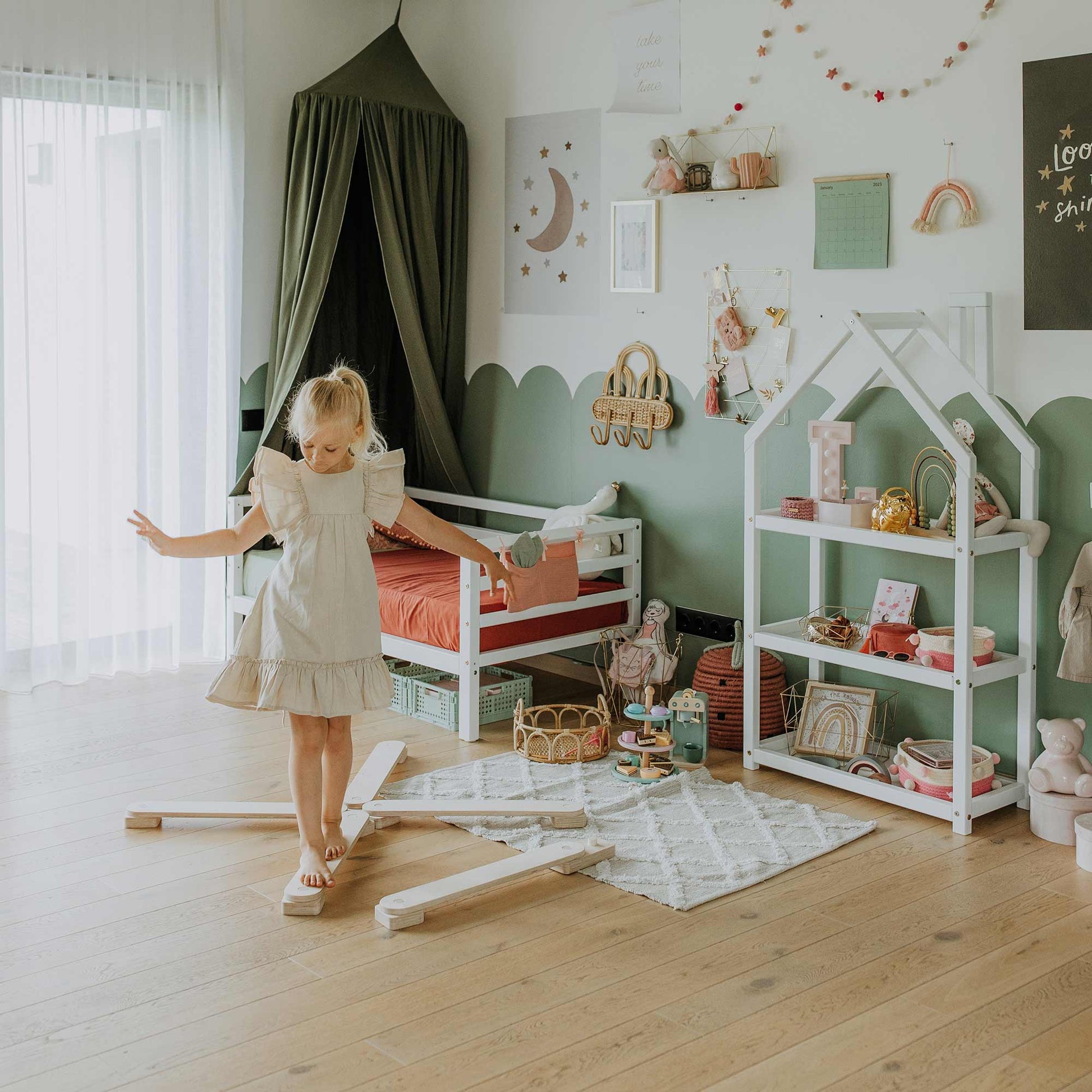 A young girl in a light dress skillfully balances on a beam from the Balance Beam Set in her child's room filled with toys, bookshelves, and Montessori-inspired decorations featuring stars, a moon, and rainbows. This playful activity beautifully enhances her motor skills.