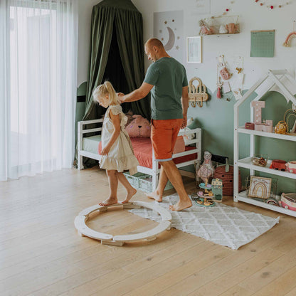 A man helps a young girl walk on the Round Balance Beams in a child's bedroom filled with toys, a canopy bed, and bookshelves. This activity toy fosters motor skills development and fun.