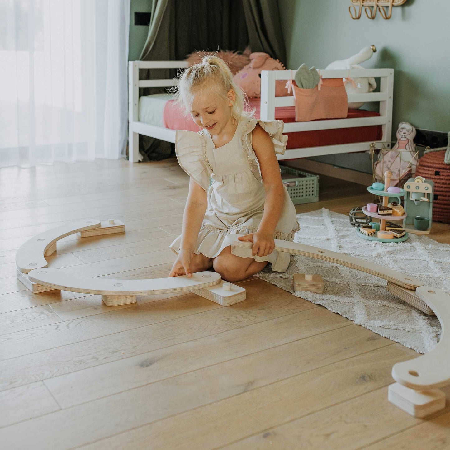 A young girl in a white dress sits on a wooden floor playing with round balance beams, enhancing her motor skills, near a white bed and toys in a cozy room.