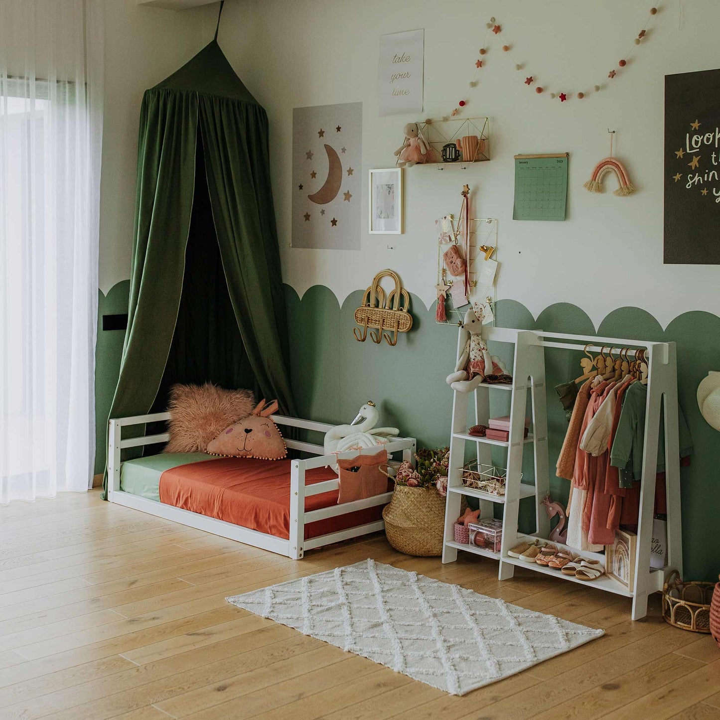 A children's room featuring a floor-level bed with a 3-sided safety rail made of solid wood, complemented by a green canopy, wall decorations, a clothing rack with dresses, a white rug, and various toys and baskets—all set against green and white walls.