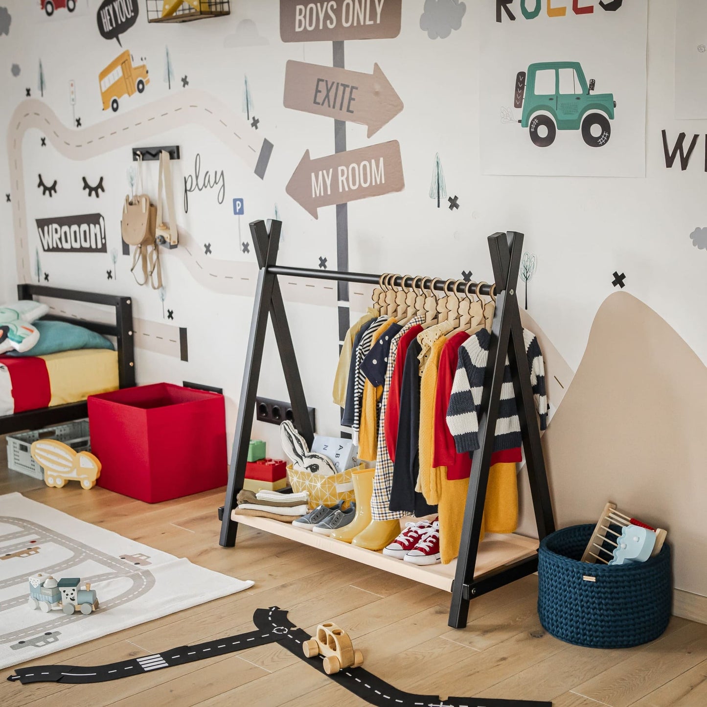 A children's room features a Kids' Clothing Rack with Storage, complete with hanging outfits and toy storage bins. The space also includes a bed and wall decorations depicting cars and adventure-themed signs. A playmat with a road pattern spans the floor, creating an organized and playful kids' storage solution.