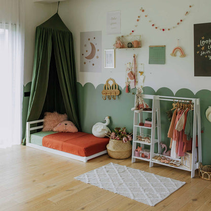 A neatly organized child's room features a small bed under a green canopy, wall decorations, a rug, and children's storage shelves made from Baltic birch plywood. A Children's wardrobe with a dress-up clothing rack displays clothes. The floor is wooden, and natural light enters from the left.