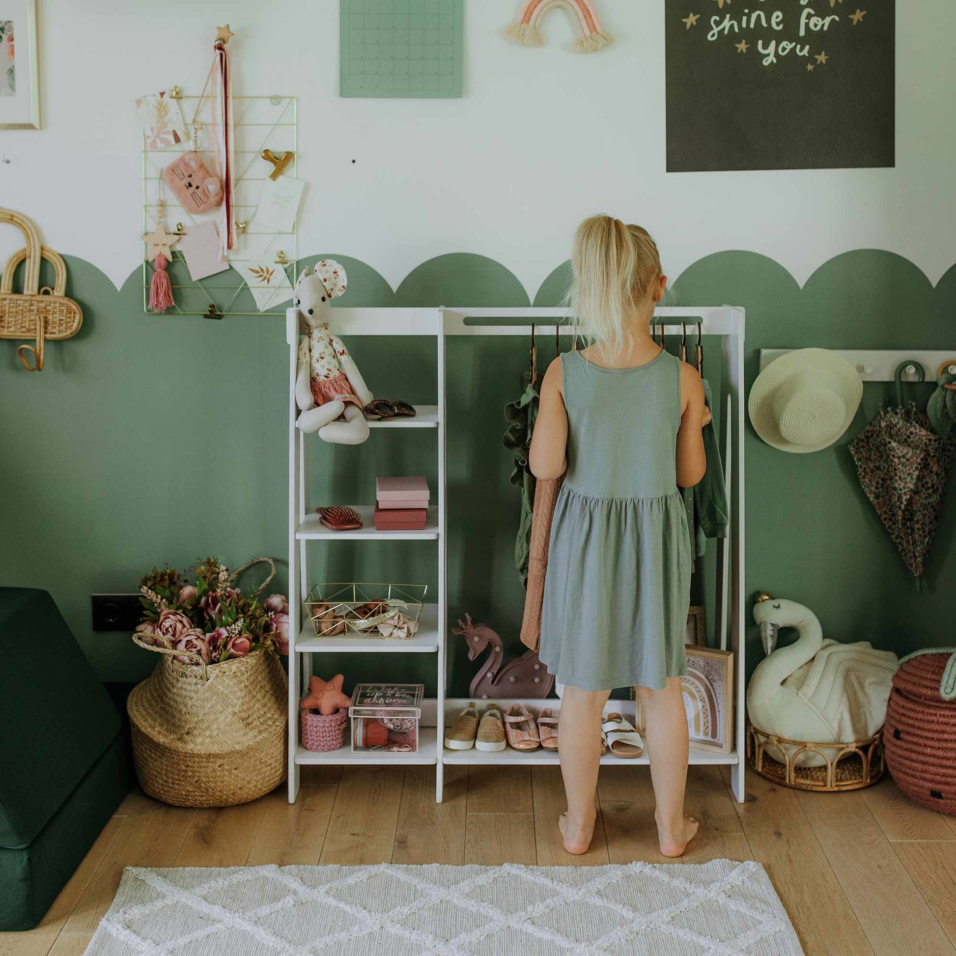 A young girl stands in front of an organized Children's wardrobe, dress up clothing rack in her bedroom, surrounded by various toys, decorations, and a rug on the floor. The room features green and white decor with a "shine for you" chalkboard and sturdy Baltic birch plywood shelves for children's storage.