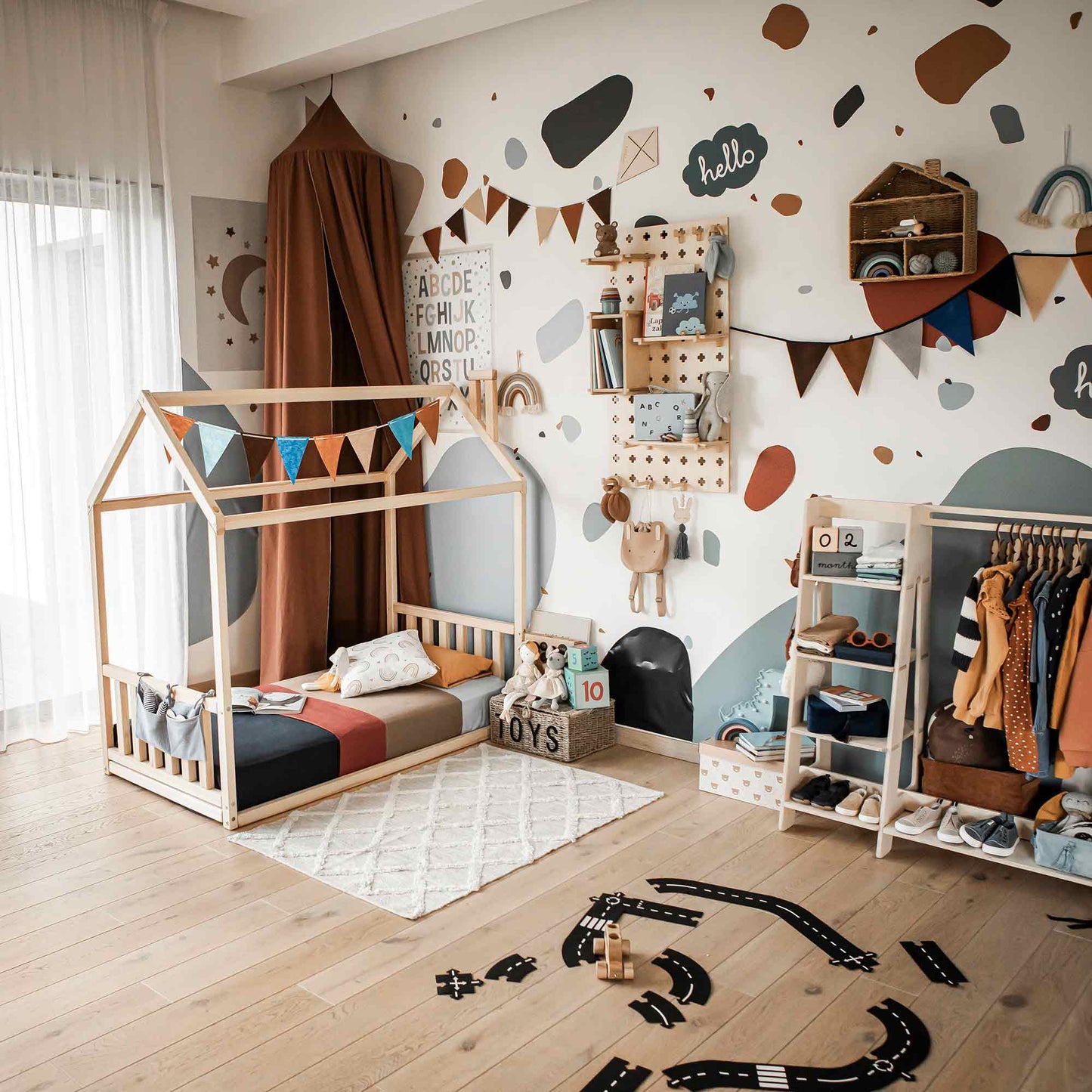 A neatly organized children's room featuring a Toddler house bed with a headboard and footboard, a shelf with toys, a clothing rack, colorful wall decals, and a play rug with a road pattern.