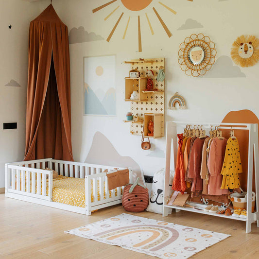 The neatly decorated children's room boasts a Montessori kids' bed with a fence, perfect for promoting independent sleeping. It also features a clothing rack adorned with colorful clothes, wall-mounted shelves, a play mat designed with a rainbow pattern, and playful wall decorations.