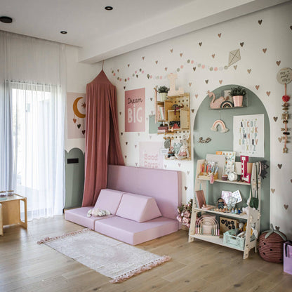 A children's playroom with pastel decor features a Montessori toy shelf brimming with toys and books, pink cushioned seating, a canopy, and decorative wall art adorned with letters and hearts. The organized space resembles a charming child's bedroom. There's a window with sheer curtains on the left.