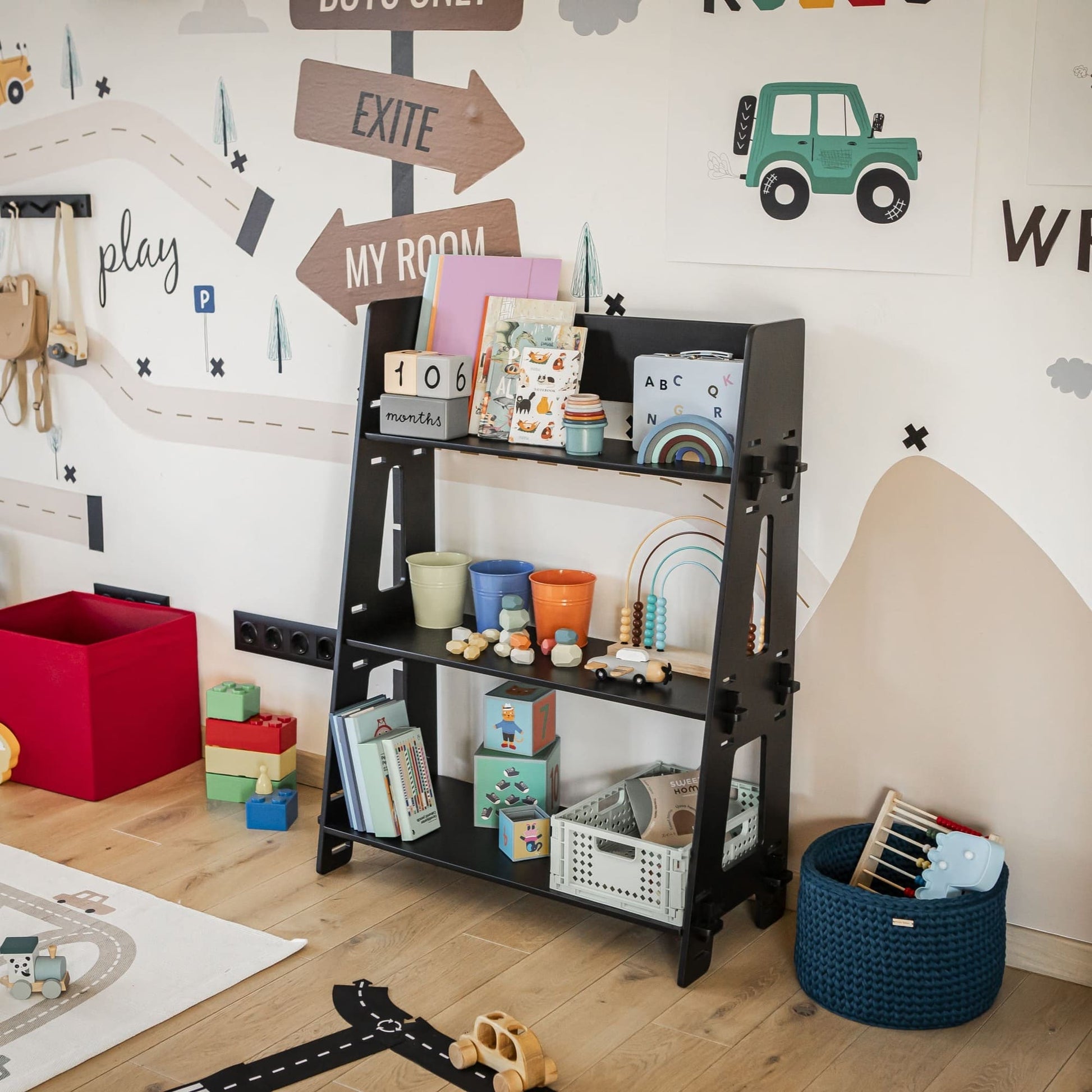 A child's bedroom efficiently serves as a playroom with an organized black Montessori toy shelf brimming with books, toy blocks, and storage bins. The walls feature a road-themed mural, while various toys are scattered across the wooden floor.