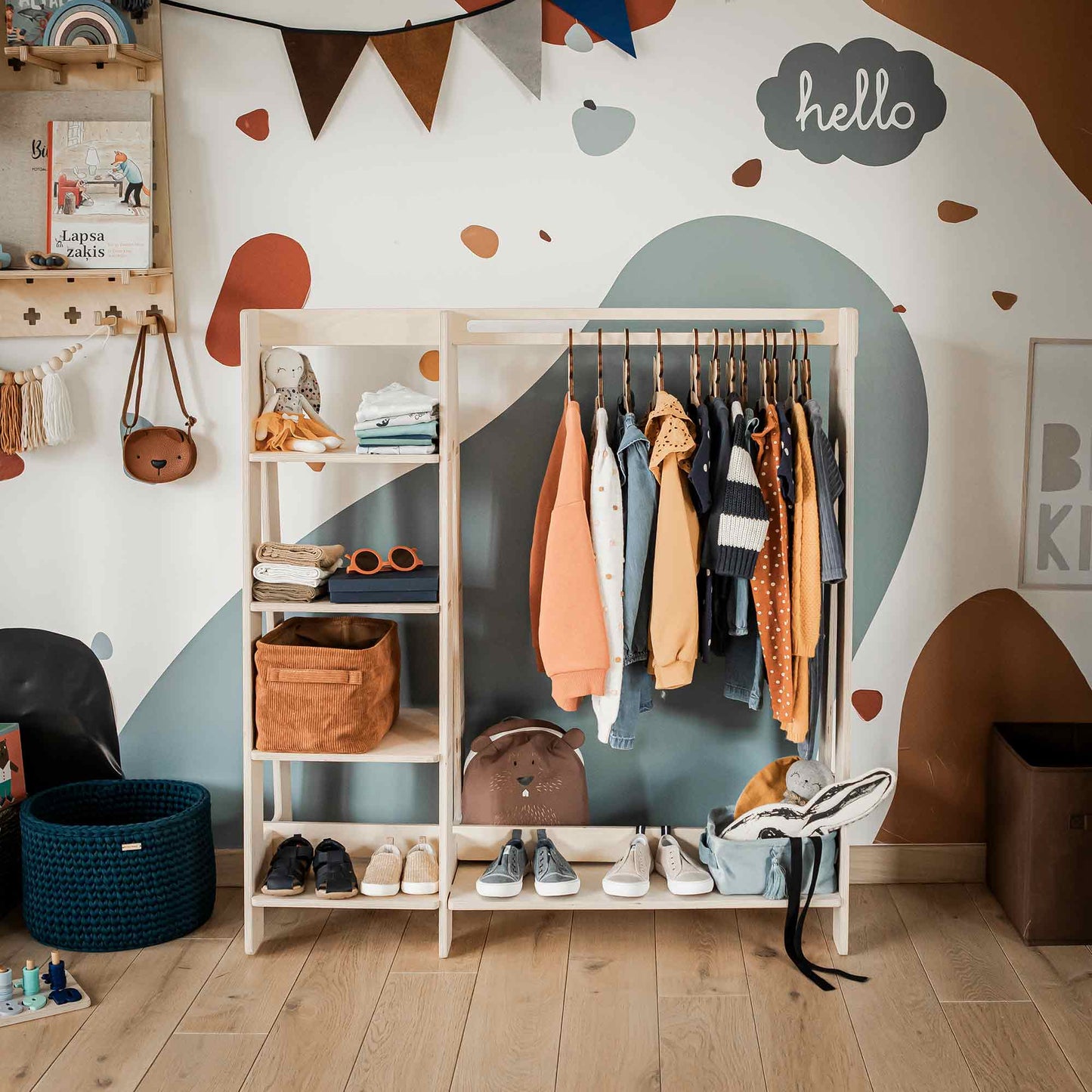 A Children's wardrobe, dress up clothing rack holds a variety of colorful clothing, accessories, and shoes. Storage shelves on the left display baskets and a clock. The background features a painted wall and decorations.