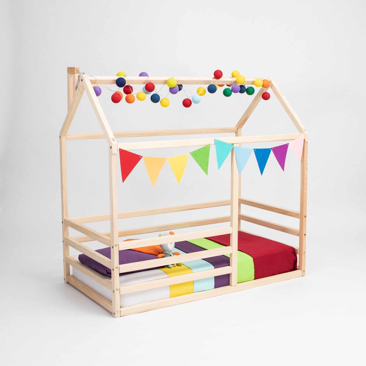 A floor level house bed with a horizontal fence, resembling a wooden house-shaped frame adorned with colorful balls and triangular banners. Inside rests a multicolored mattress and pillows, making it the perfect preschool bed for imagination and comfort.