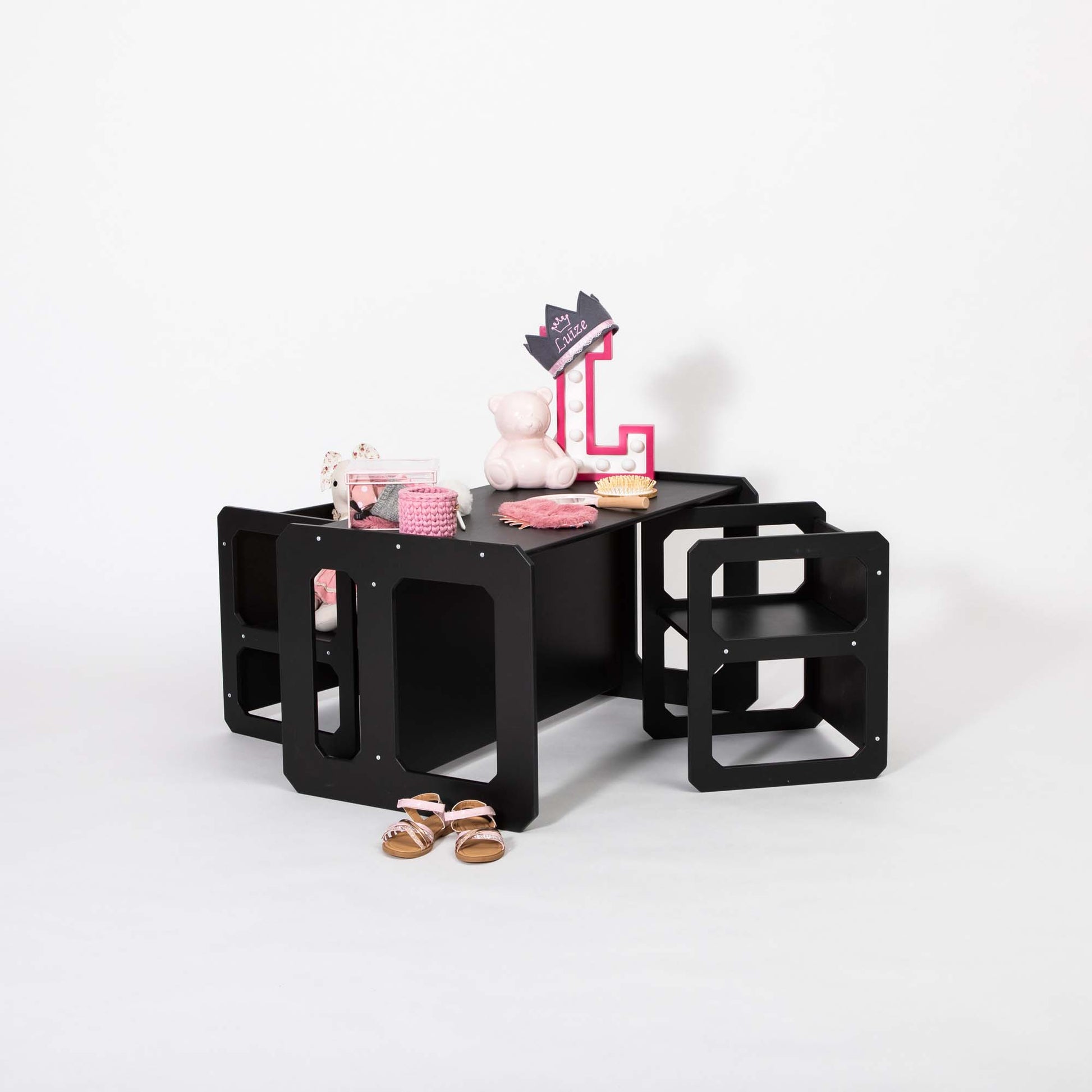 The Montessori weaning table and 2 chair set, black and modular, holds a variety of items including a teddy bear, children's shoes, a crown sign, and a small container of pink items. This setup is perfect for fostering toddler independence and fine motor skill development.
