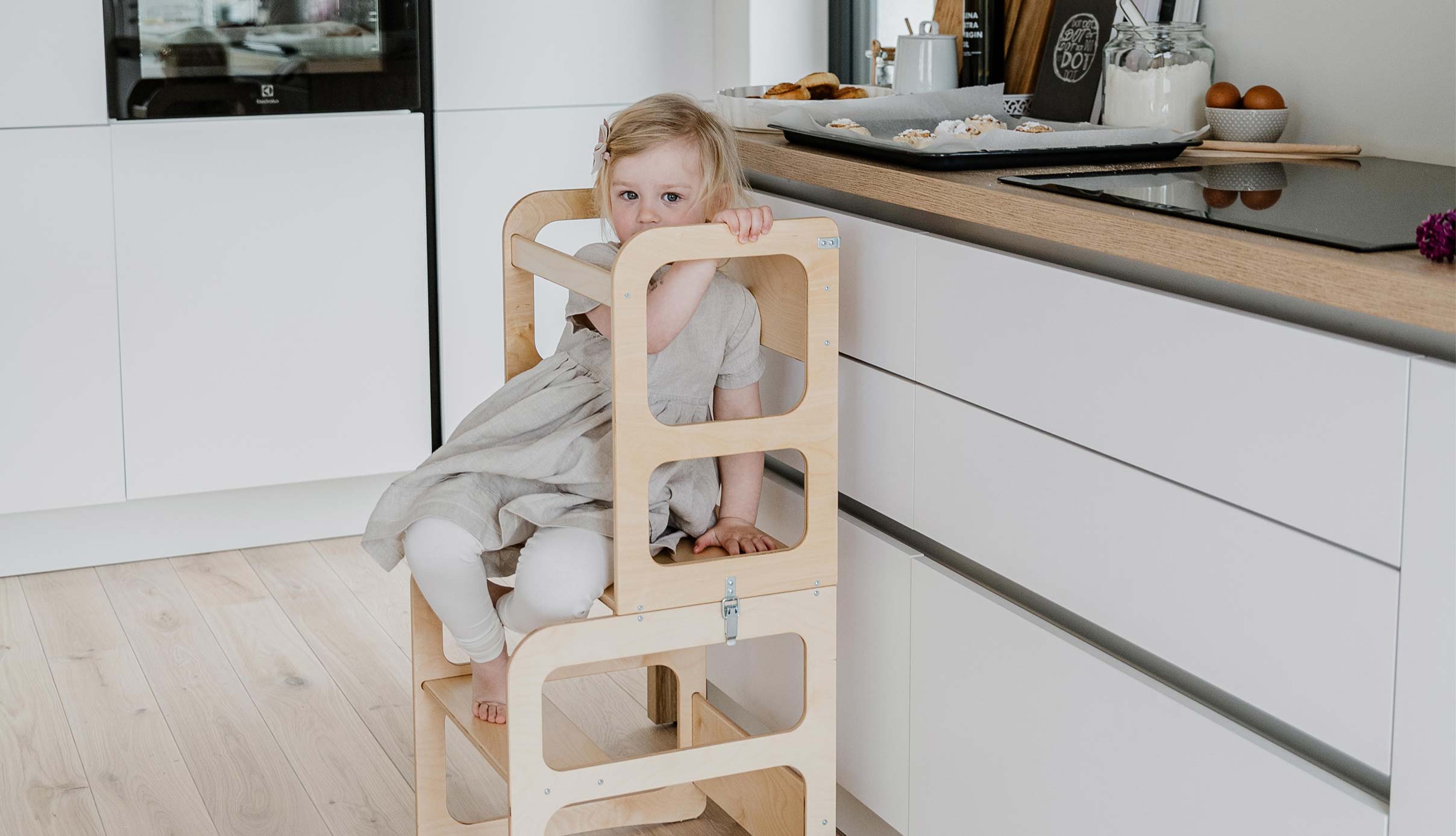A little girl sitting on a wooden high chair in a kitchen.