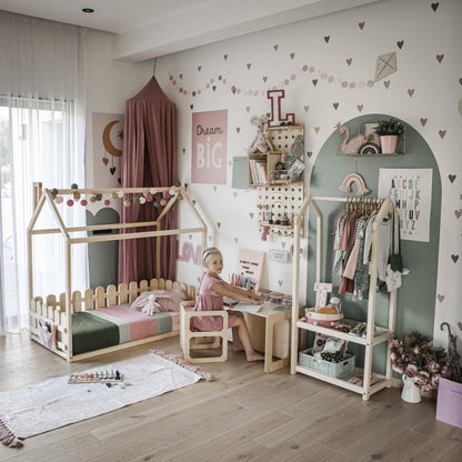 A child sits at a Montessori weaning table and chair set in a playfully decorated children's room featuring a house-shaped bed, toys, shelves with books, clothes racks, and wall decorations that foster toddler independence and fine motor skill development.