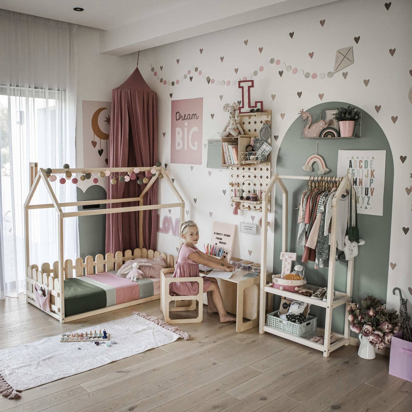 A child sits at a small desk in a well-organized, pastel-colored bedroom with a canopy bed, wall shelves, and decorative items including heart decals and alphabet posters. The Montessori weaning table and 2 chair set not only enhances fine motor skill development but also fosters independence.