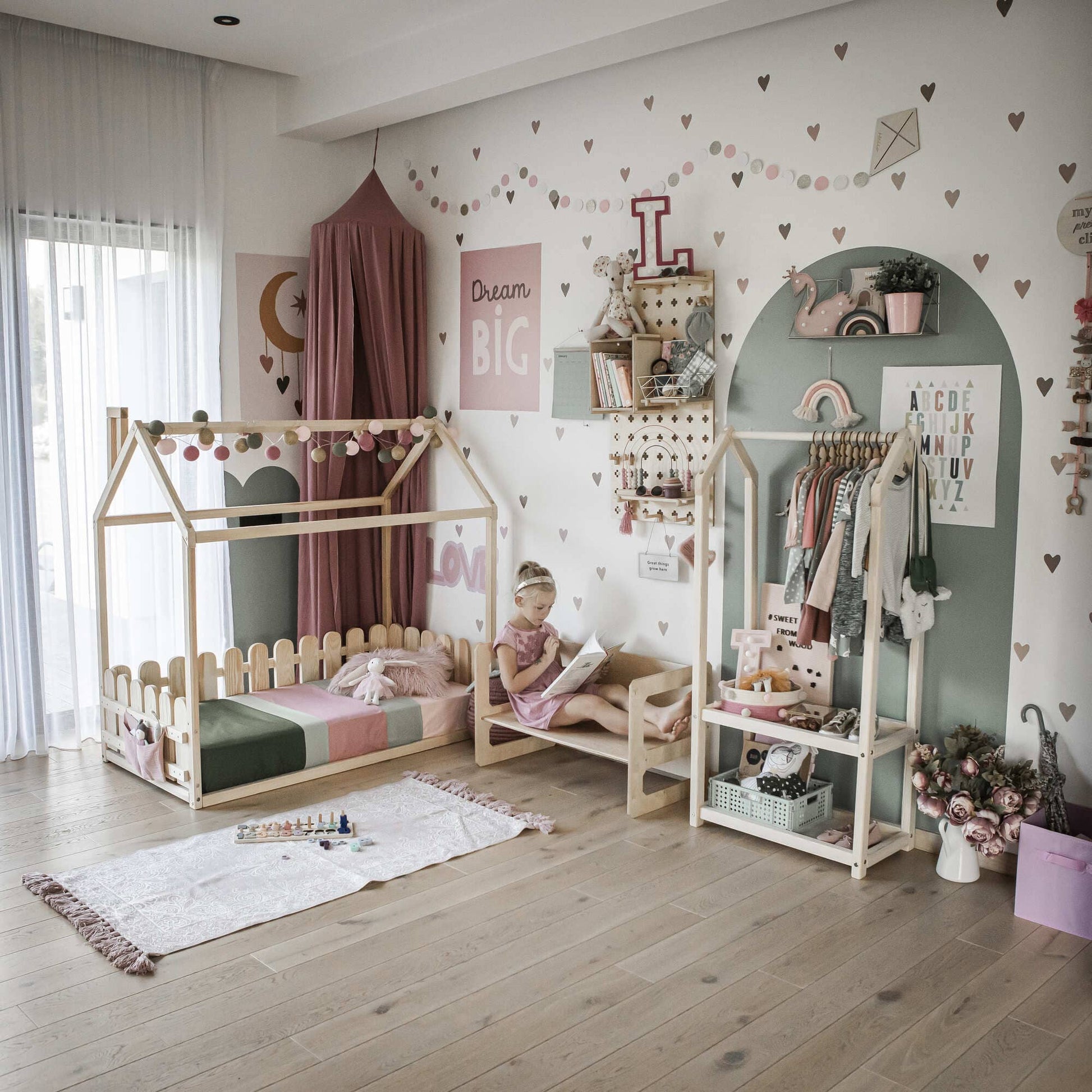 A children's room with pastel decor, featuring a canopy bed, a girl reading on a chair, shelves with toys and books, and a clothing rack. Walls have heart and text stickers. A rug and toys are on the floor next to a Montessori weaning table designed for fine motor skill development.