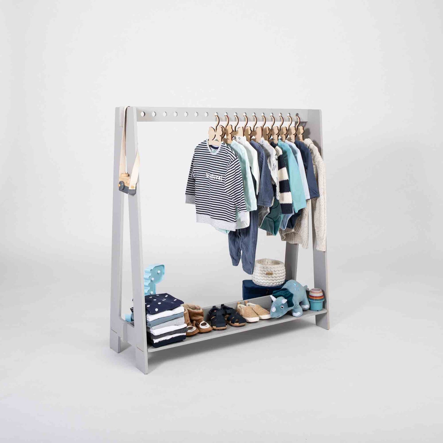 A Montessori clothing rack designed for children, featuring several hangers to hold a variety of shirts and sweaters, along with shelves below for neatly organizing folded clothes, shoes, and a plush toy.