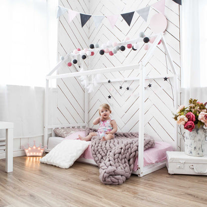 A little girl is sitting on a Sweet Home From Wood house-frame bed with a headboard in a pink and white room.