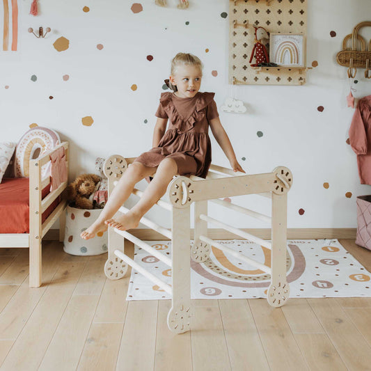 A Montessori-inspired little girl sitting on a wooden bed in a room with polka dots, engaging with her 2-in-1 Climbing cube/ table and chair + Transformable climber + a ramp.