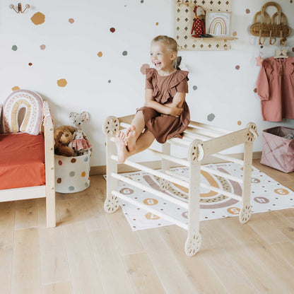A little girl sitting on a wooden chair surrounded by a Climbing triangle, Transformable climbing gym, and a ramp in a child's room.