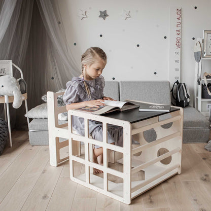 A little girl is reading a book in a room with a wooden desk and an indoor Transformable climbing cube / table and chair + ramp.